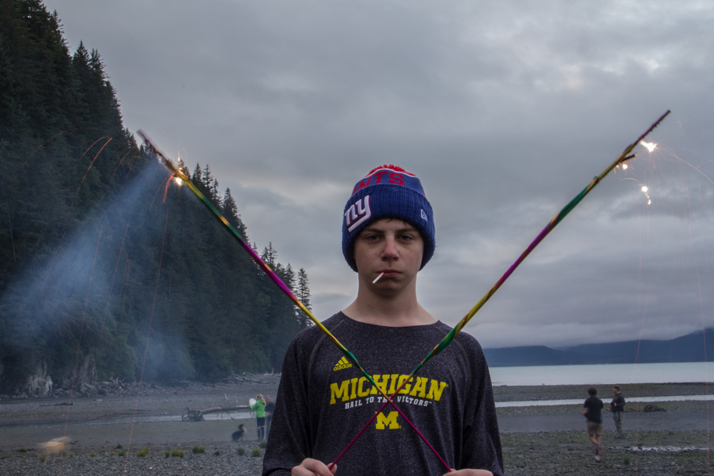 Jakob Fox, for his first time in Alaska on the 4th of July stands by the water holding to sparklers in resurrection bay. He stands by the water with a lollipop after hiking to Tonsina Point along the canines head trail.