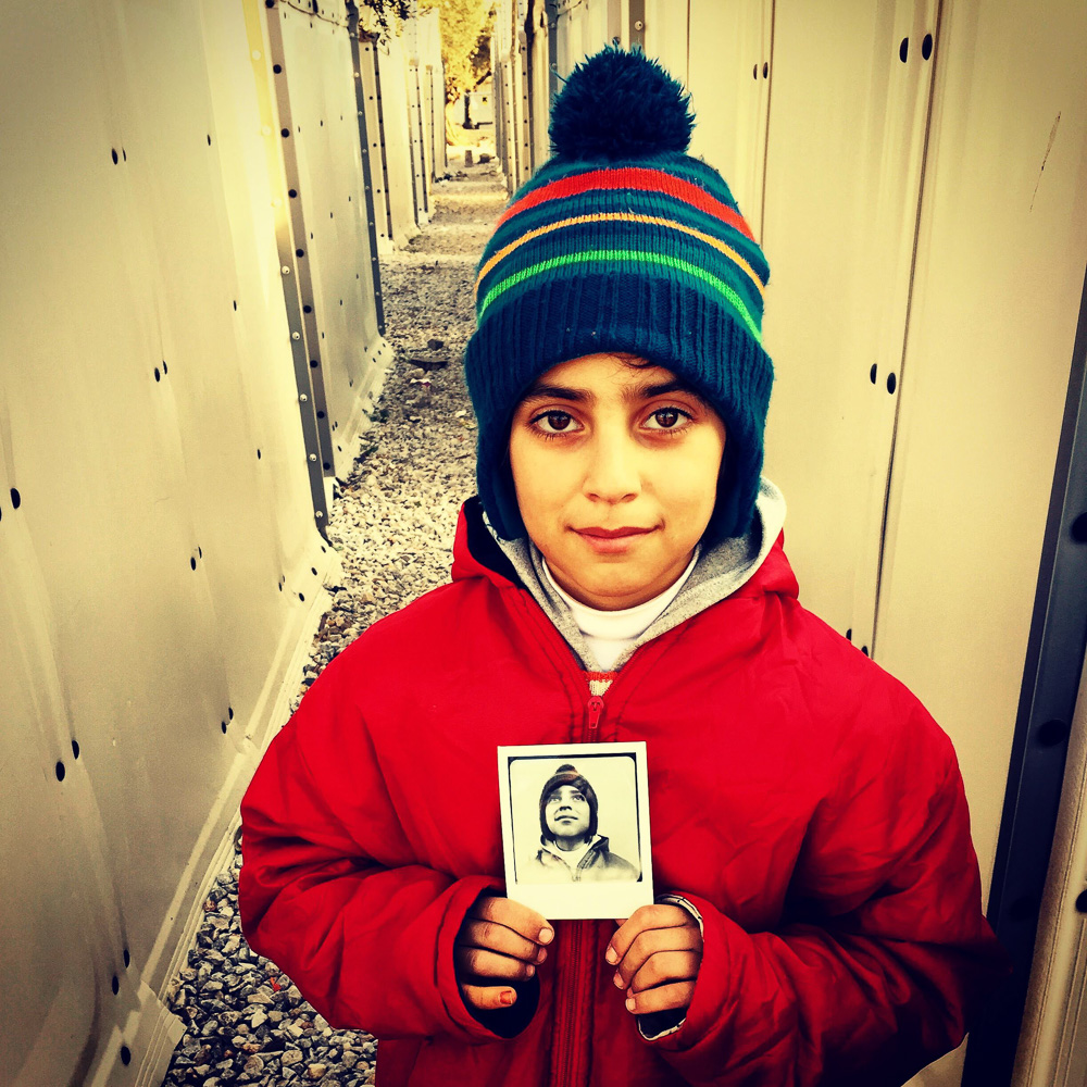 © Wayne Martin Belger, After "Us & Them" photo shoot in the Syrian refugee camp, Kara Tepe in Lesbos, Greece. They young Syrian boy had just been photographed by the "Us & Them" camera. He's holding a 4" x 5" Polaroid given to him by Wayne.