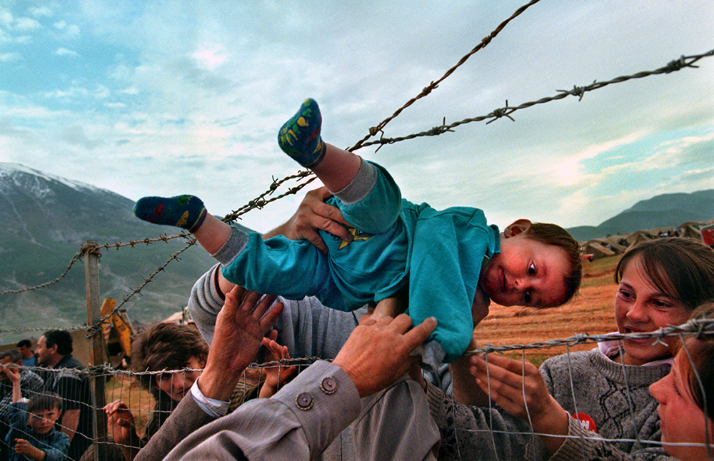 05/03/1999 - SLUG: FO/LIFE IN EXILE - THE CAMPS DATE: 5/3/99 - PHOTOG: Carol Guzy/The Washington Post  -  LOCATION: Kukes, Albania. CAPTION: LIFE IN EXILE - THE CAMPS:  Agim Shala, 2 years old, is passed thru the barbed wire fence at the Arab camp as members of the Shala family are reunited after fleeing Kosovo.  The relatives who just arrived from Prizren had to stay outside the camp until shelter was available.