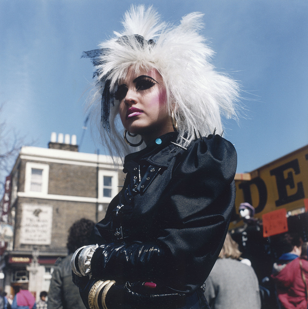 A stylish young lady in punk attire of black satin dress with spiked shoulder pads, wild white wig, heavy make-up and gold and silver bangles, stands on a Camden street as shoppers hunt for bargains in the market behind. Photograph by Shirley Baker  *EDITORIAL USE ONLY - PICTURE MUST ONLY BE REPRODUCED IN RELATION TO ITS ORIGINAL CONTEXT / SUBJECT MATTER*     Date: 1986