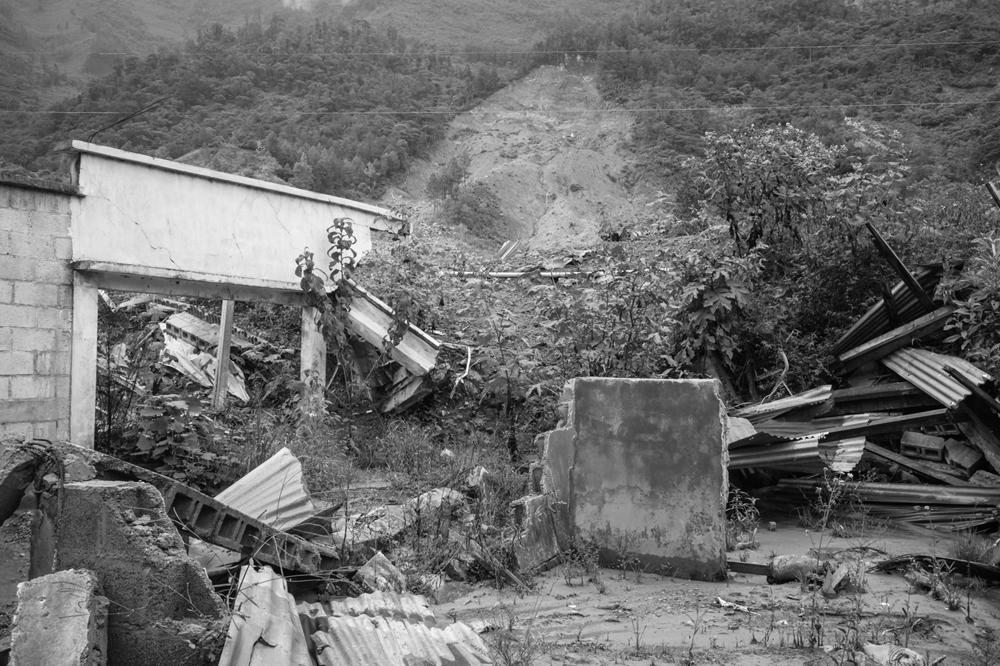 View of the destruction left behind by the landslide, with a crumbled steel, stucco, and cement block structure in the foreground, and the mountain responsible for the disaster looming in the background, serves as a reminder of the devastating event that wiped out the village of Queja.
