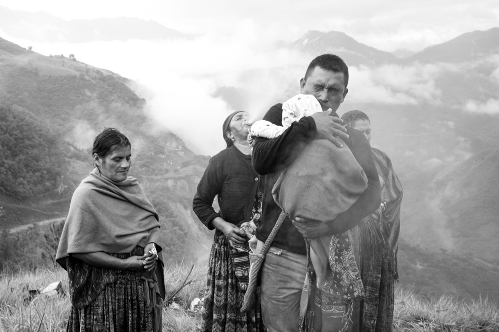 At the time of the landslide, Jorge's wife, Sonia, was pregnant and at home with their other children while Jorge was at work. Here, Jorge holds his baby while the village elder leading the Mayan ceremony blows rum out of her mouth at the back of his head as part of one of the rituals performed during the event.