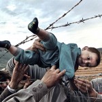 KOSOVO'S SORROW - ETHNIC CLEANSING
"REUNION"
Kosovar refugee Agim Shala, 2 years old, is passed through the barbed wire fence into the hands of grandparents at the camp run by United Arab Emirates in Kukes, Albania. The members of the large Shala family were reunited here after fleeing Prizren in Kosovo during the conflict. . (The grandparents had just crossed the border at Morina). The relatives who just arrived had to stay outside the camp until shelter was available. The next day members of the family had tents inside. The fence was the scene of many reunions. When the peace agreement was signed, they returned to Prizren to find their homes only mildly damaged. There were tears of joy and sadness from the family as the children were passed through the fence, symbolic of the innocence and horror of the conflict. (Photo by Carol Guzy/The Washington Post) Kukes, Albania May 3, 1999