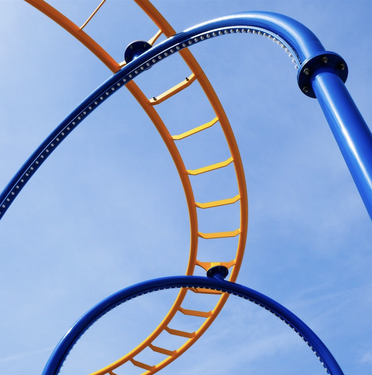     Curving blue and orange roller-coaster tracks loop across a soft blue sky in a bold geometric composition. ©CynthiaGladis