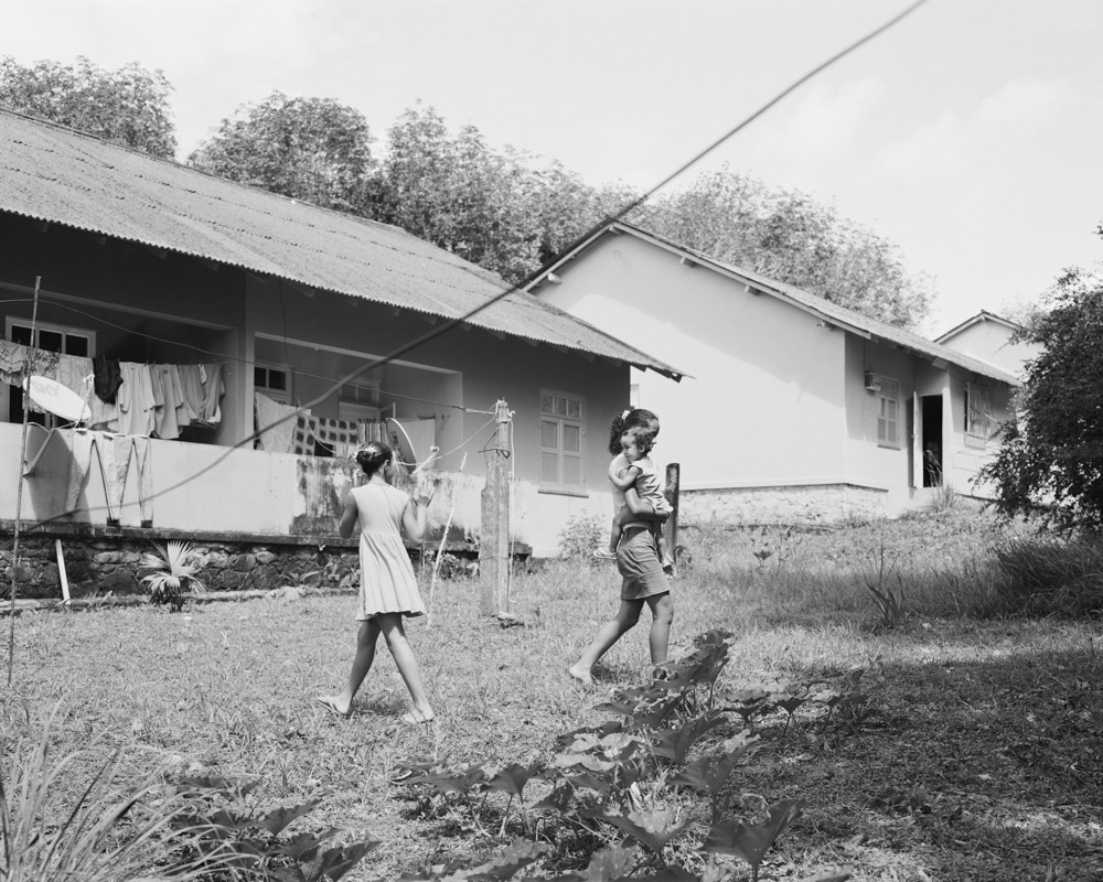 Sisters Ivna and Ingrid Souza Playing with Cousin Monick Farias in the Family Backyard with Squash Plants in the Foreground at Company Housing Complex Vila 4, Michelin Rubber Plantation, Bahia, Brazil. Archival pigment print, 32 × 40 in. © Karolina Karlic. Overshoot with Yogan Muller.